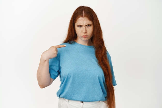 Boos mokkend tienermeisje met rood lang haar, fronsend overstuur, wijzend met de vinger naar zichzelf en klagend, ontevreden starend, staande tegen een witte achtergrond