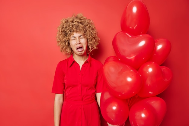 Boos ontevredenheid krullend haired jonge vrouw draagt stijlvolle jurk voelt neerslachtig kreten van wanhoop eenzaam en onbemind op Valentijnsdag draagt jurk geïsoleerd op rode achtergrond Zwart-wit schot