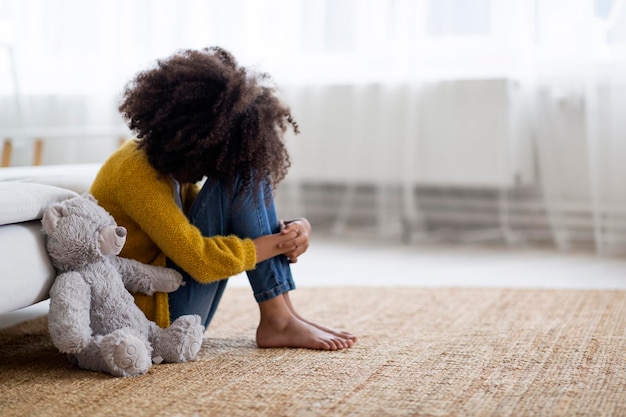 Boos preteen zwart meisje zittend op de vloer met teddybeer