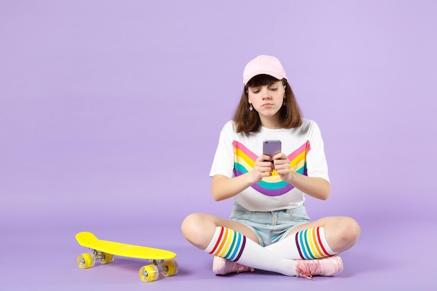 Boos tienermeisje in levendige kleding zitten in de buurt van skateboard, met behulp van mobiele telefoon, sms-bericht geïsoleerd op violet pastel achtergrond te typen. Mensen oprechte emoties, lifestyle concept. Bespotten kopie ruimte.