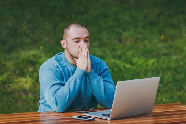 Boos verdrietig man zakenman of student in casual blauw shirt, zittend aan tafel in stadspark met behulp van laptop buitenshuis werken, hand in hand op zijn gezicht, bezorgd over problemen. mobiel kantoorconcept.