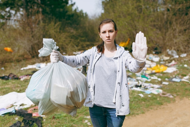 Boos vrouw in vrijetijdskleding die vuilniszakken vasthoudt en stopgebaar toont met palm in bezaaid park