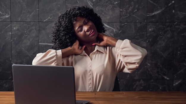 Bored afro-american secretary with curly hair stretches back\
sitting at brown wooden table with grey laptop by marble wall in\
empty office