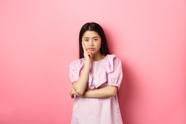 Bored asian teen girl look indifferent at camera, lean face on hand in skeptical pose, standing reluctant against pink background.