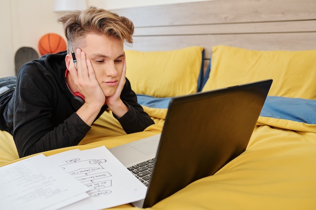 Foto studente universitario annoiato sdraiato sul letto, guardando lezioni online sulla programmazione e disegnando diagrammi di flusso in quaderno