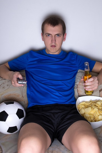 Bored man in football uniform sitting in living room and watching tv with beer and chips