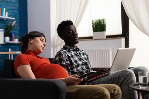 Moglie incinta annoiata che cambia programmi televisivi mentre il marito è seduto sul divano con il laptop in grembo. le coppie multietniche incinte amano stare insieme mentre guardano la televisione e usano il computer.