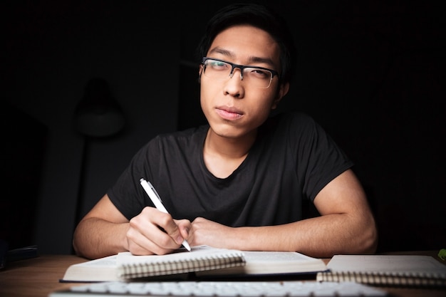 Bored serious asian young man in glasses learning and writing in the evening at home