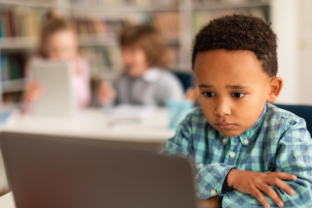 Bored serious black kid doing homework or watching video on laptop computer sitting at desk in