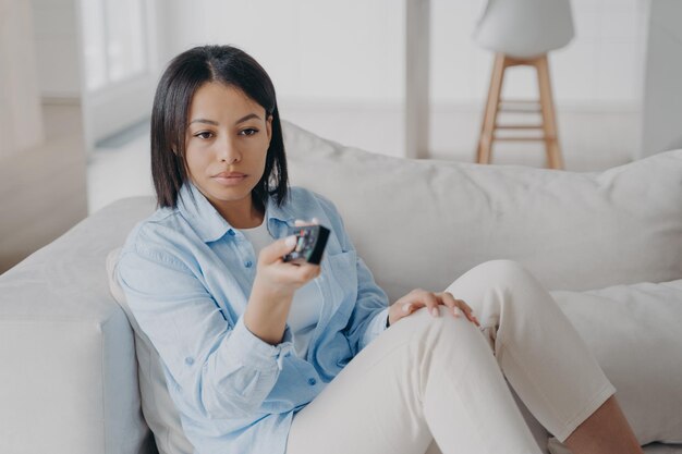 Bored spanish woman holding remote control and watching bad movie on tv and shifts the channels