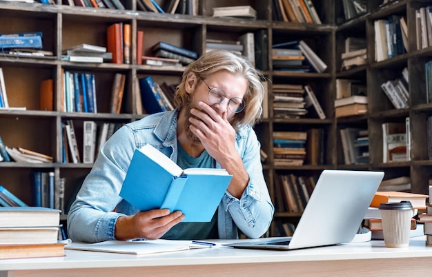 Bored tired guy student yawning reading book studying in library