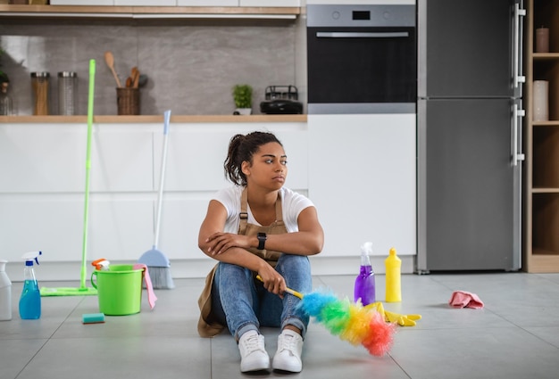 Bored tired millennial black housewife sit on floor with cleaning supplies and brush looking at empty space