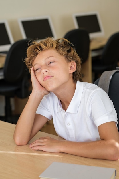 Bored and tired schoolkid leaning on hand while sitting at table in classroom during lesson at school