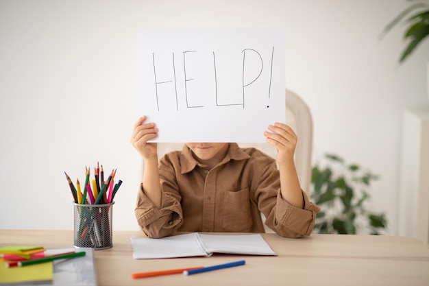 Bored upset tired caucasian little child sits at table shows paper with inscription help suffer from overwork