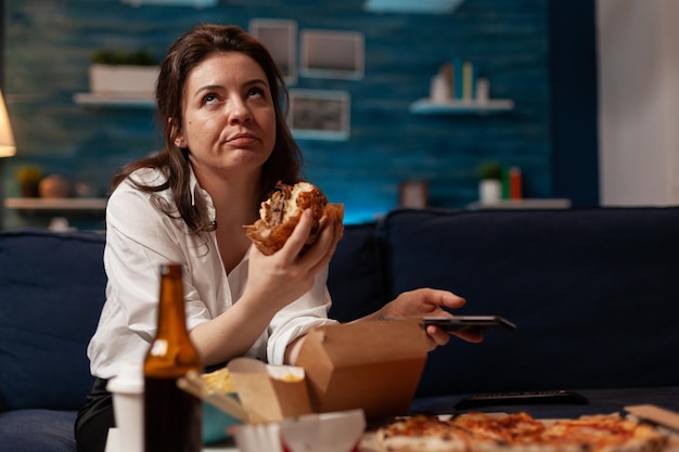 Bored woman browsing social media on smartphone eating\
delicious burger takeout dinner sitting on sofa in home living\
room. caucasian female having takeaway dinner junk-food order.