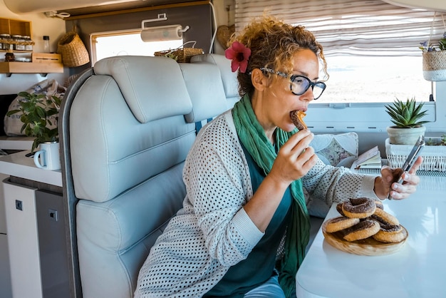 Photo bored woman eating a lot of donuts sitting at the table inside a camper van during travel lifestyle female people enjoy vacation in motorhome having relax and using mobile phone and connection