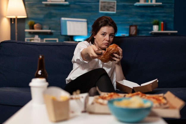 Photo bored woman sitting on sofa eating tasty delicious burger holding remote switching channels on television in living room. tired person browsing tv stations while eating takeaway hamburger.