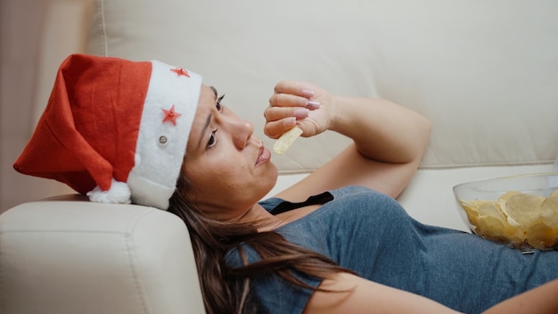 Bored woman wearing santa hat and eating chips from bowl