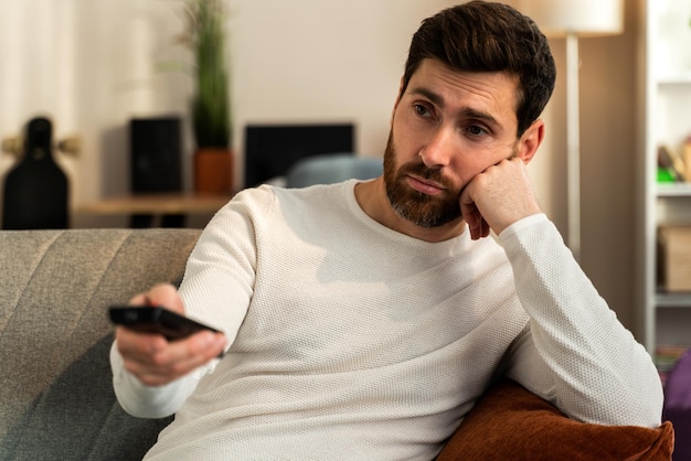 Giovane uomo caucasico annoiato che tiene il telecomando della tv mentre è seduto su un divano a casa mentre si rilassa durante il fine settimana