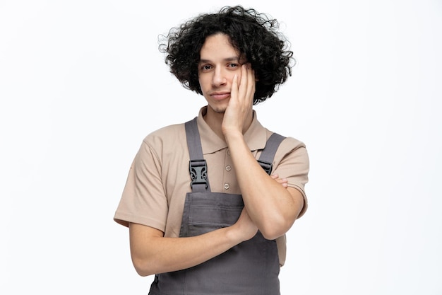 Bored young male construction worker wearing uniform keeping hand on face looking at camera isolated on white background