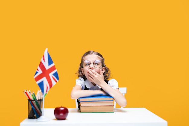 Boring caucasian schoolgirl sits at the desk with stack of books english lesson great britain flag