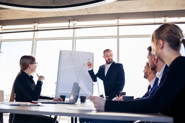 The boss stands near the board with graphs, demonstrates statistics, various personnel attending the training, introduces the new products of the company, reports on the results of work for partners.