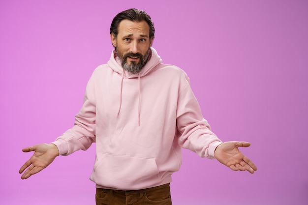 Bothered confused mature adult bearded man grey hair in pink hoodie arguing look pissed offended shrugging spread hands sideways dismay look clueless camera asking why, purple background.