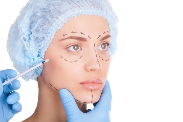 Botox injection. Beautiful young woman in medical headwear and sketches on face looking away while doctors hand making an injection in face
