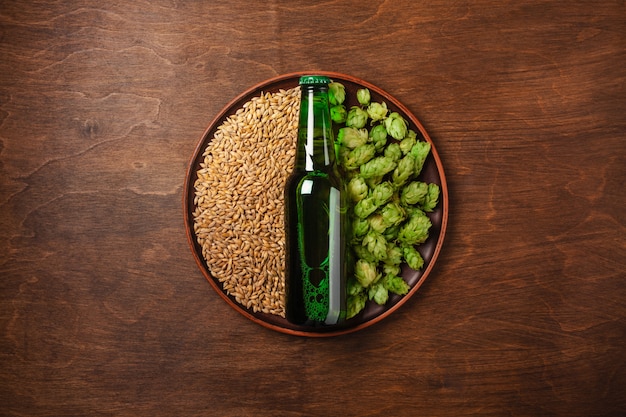 A bottle of beer on a green fresh hop and wheat grain in a plate against the wooden brown board