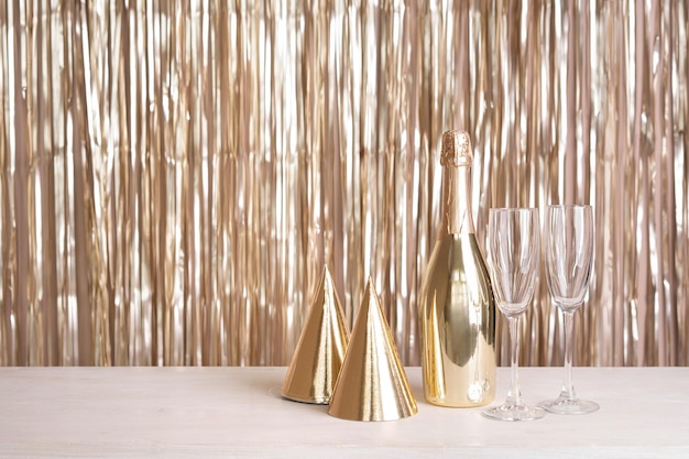 A bottle of champagne glasses and party hats are on the table against the backdrop of a golden tinsel curtain