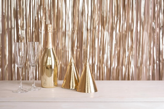 A bottle of champagne glasses and party hats are on the table against the backdrop of a golden tinsel curtain