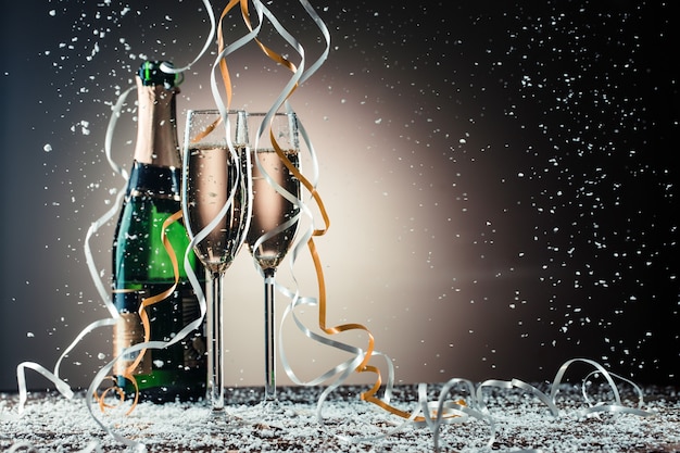 Bottle of champagne, wine glasses with silver and golden ribbons, snowfall on dark background with backlight. Holiday concept