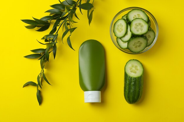 Bottle of cosmetics, cucumber slices and branch on yellow surface