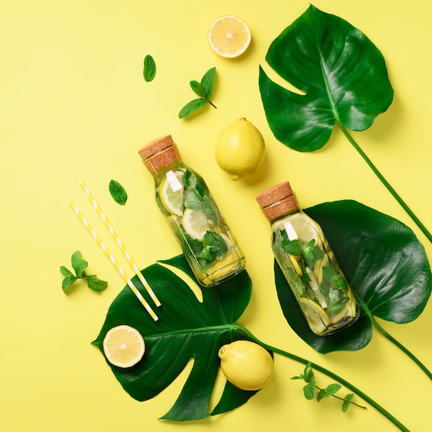 Bottle of detox water with mint, lemon and tropical monstera leaves on yellow background. 