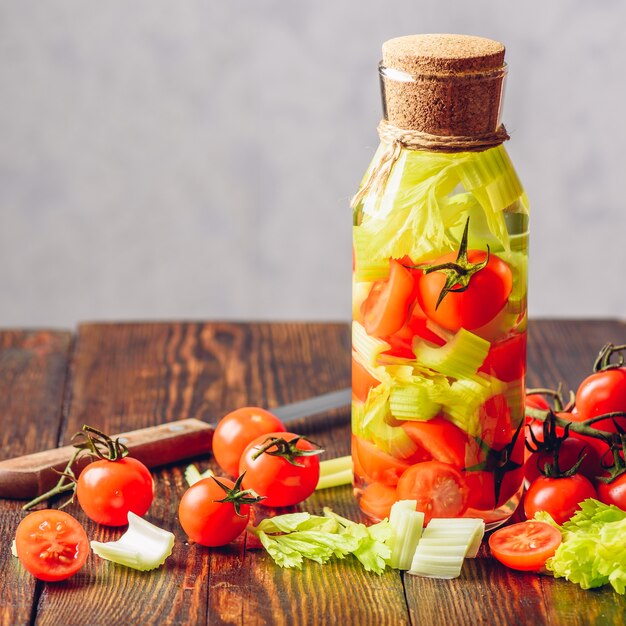 Bottiglia di acqua aromatizzata con verdure: pomodorini e gambi di sedano. ingredienti e coltello sulla tavola di legno. copia spazio.