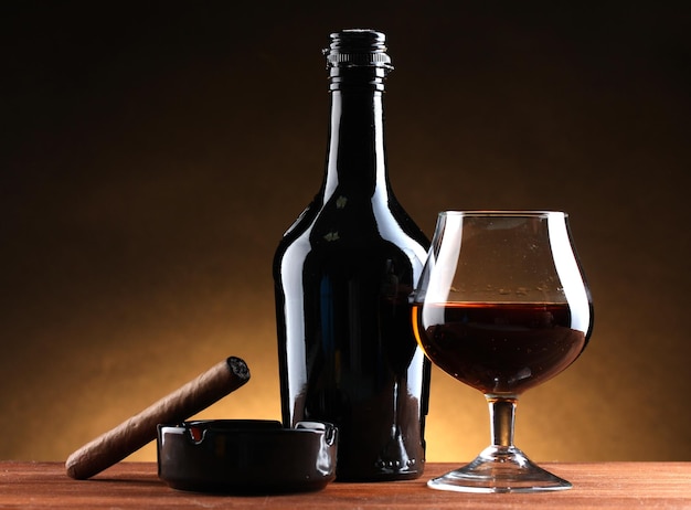 Bottle and glass of brandy and cigar on wooden table on brown background