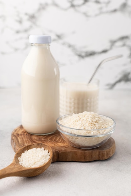 A bottle and a glass of rice milk with a bowl of rice and a wooden spoon on a wooden board on a light background