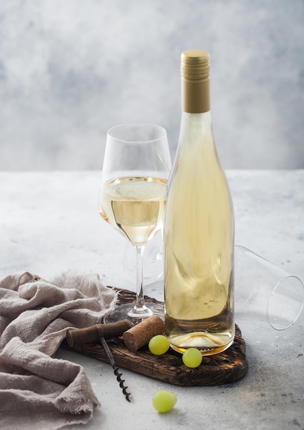 Bottle and glass of white homemade wine with corks corkscrew and grapes on wooden board with linen cloth on light table background