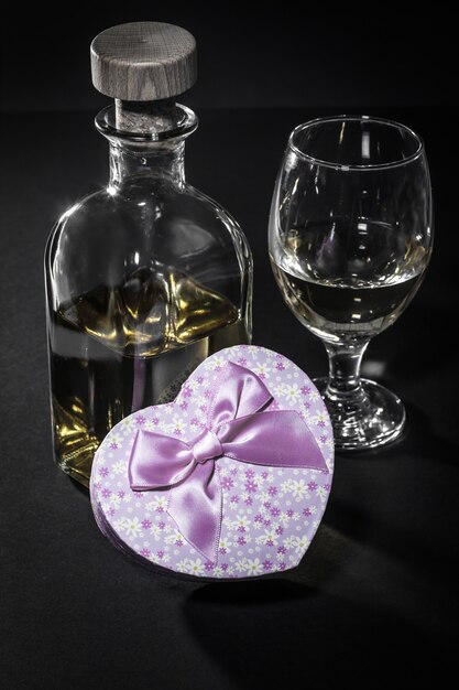 Bottle and glass of white wine, gift box in shape of a heart on black background. Valentine's day greeting card concept.