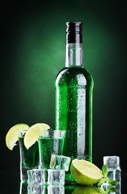 Bottle and glasses of absinthe with lime and ice on green background