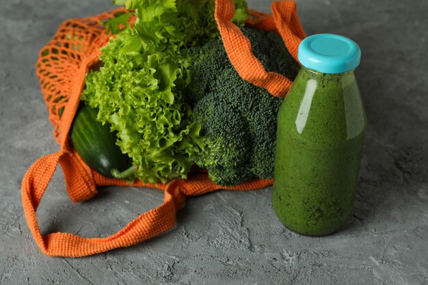 Bottle of green smoothie and straw bag with ingredients on gray textured background
