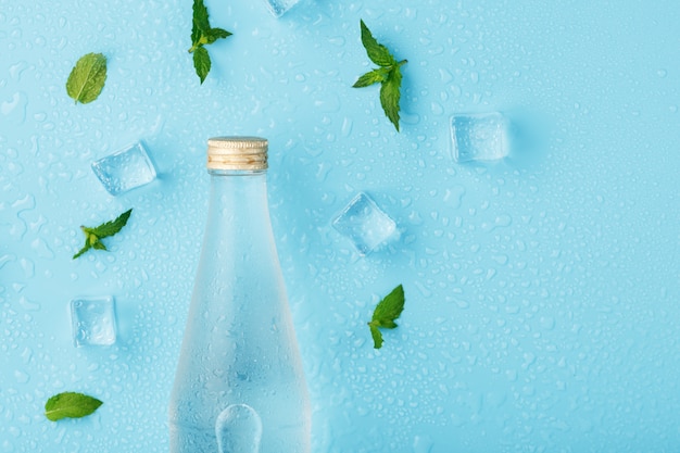 Una bottiglia di acqua ghiacciata, cubetti di ghiaccio, gocce e foglie di menta sul blu.