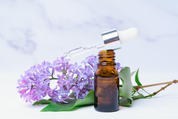 A bottle of lilac essential oil and a branch with flowers on a white marble background