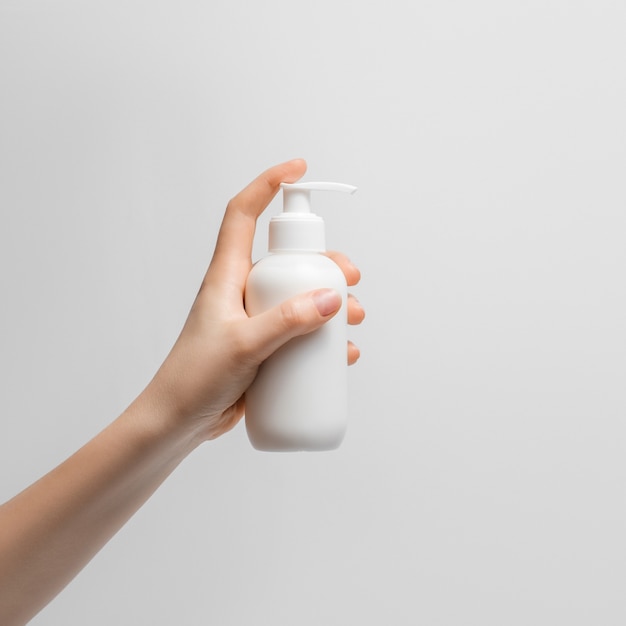 Бутылка жидкого крема для рук в женской руке на светлом фоне. Белая бутылка с дозатором, ухоженные руки, натуральные короткие ногти.