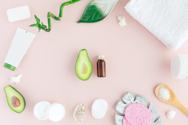 Bottle of oil, green leaves and bamboo, white towel and fresh avocado on pink, flatlay