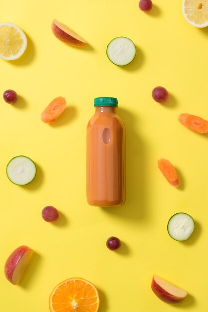 Photo bottle of orange fruit and vegetable juice in the center of the image surrounded by pieces of fruits and vegetables of various colors on a yellow background