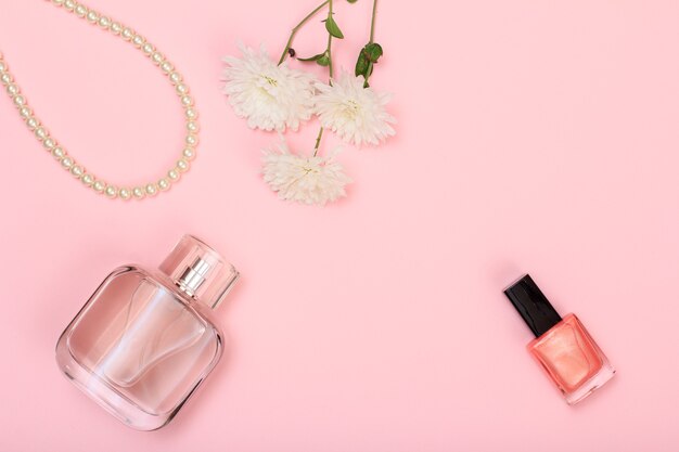 Bottle of perfume, beads, bottle with nail polish and flowers on a pink background. Women cosmetics and accessories. Top view.