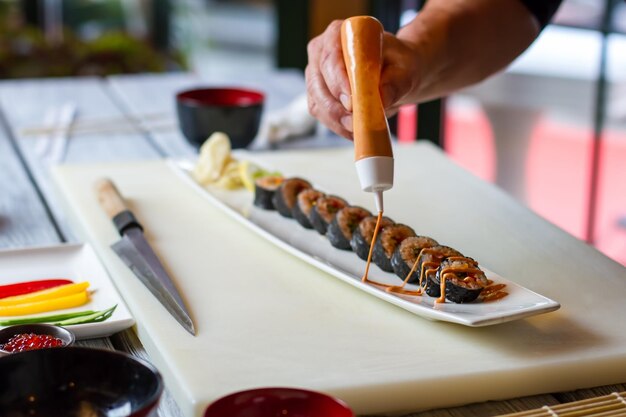 Foto la bottiglia versa la salsa sul sushi. mano d'uomo con bottiglia a pressione. salsa dolce per involtini futomaki. piatto giapponese cucinato dallo chef.