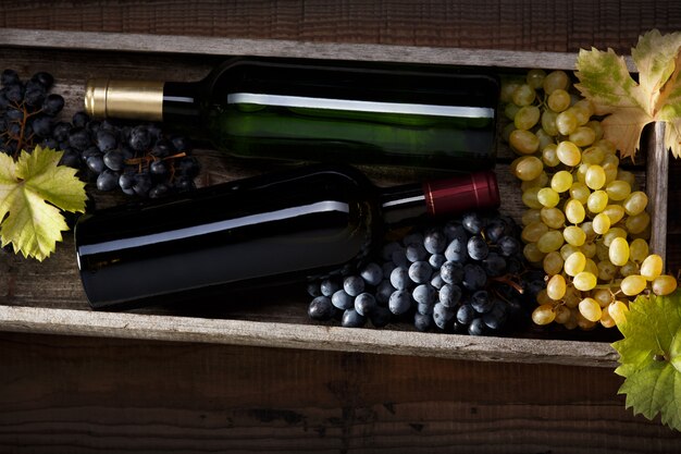 bottle of red wine and a bottle of white wine, black grapes and white grapes with grape leaves on an old wooden table.