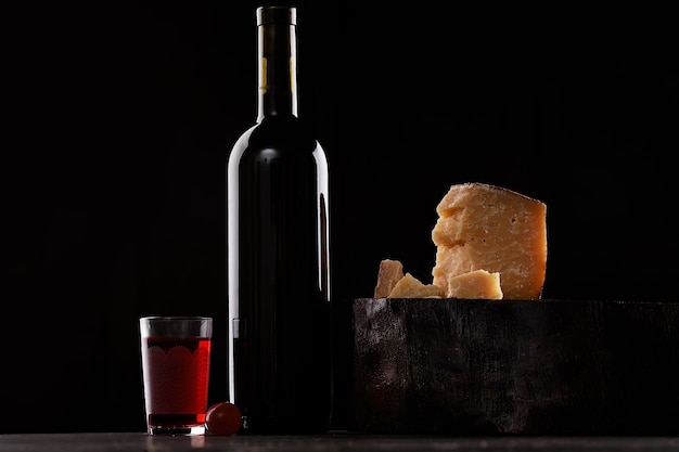 A bottle of red wine and a glass of red wine, an expensive sort of cheese with mold and grapes. On black background. Place for logo.